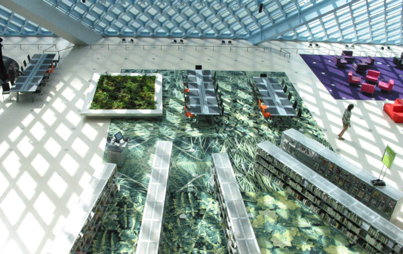 A modern library interior with patterned floors, bookshelves, tables, a green plant display, and scattered seating areas, viewed from above—perfect inspiration for your Friday 5 design ideas.