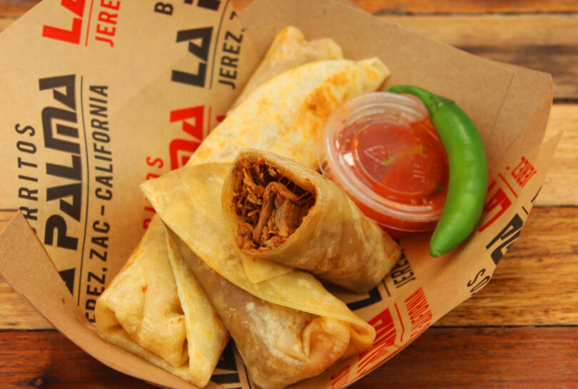 Three rolled tacos filled with shredded meat, a side of red salsa in a plastic cup, and a whole green chili pepper—all served on branded paper in a cardboard tray. Perfect for enjoying with the Friday 5 special!.