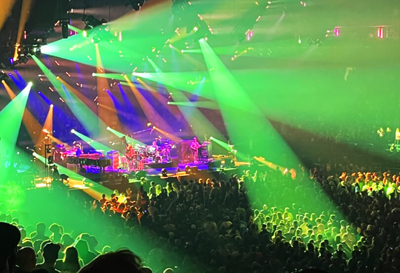A band performs on stage under vibrant green and yellow lights during the Friday 5 event, with a large crowd of people watching in an indoor concert venue.