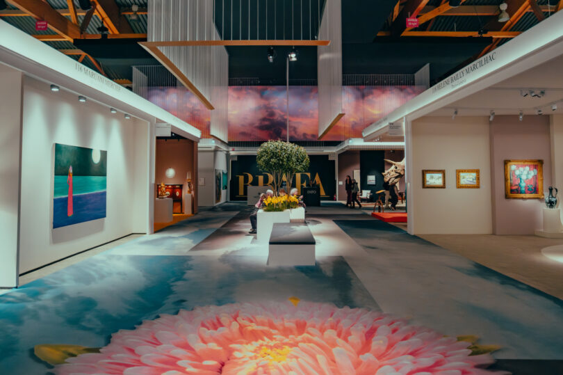 A spacious art gallery with paintings on white walls, a central green plant display, and a large pink flower image on the floor under bright lighting—perfect for a Friday 5 visit.