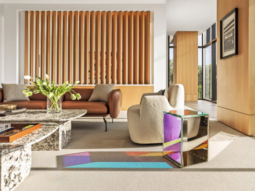 Modern living room with a brown sofa, beige armchair, marble coffee table, and glass side table casting colorful reflections—perfect for a Friday 5 gathering—plus a wooden vertical slat divider in bright natural light.