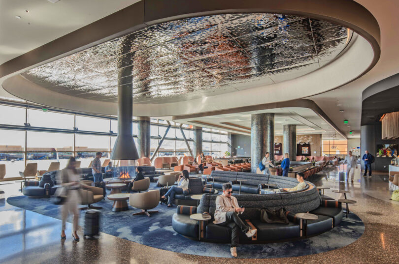 Spacious airport lounge with modern seating, a central fireplace, and several people relaxing or walking with luggage. Large windows provide natural light, creating an inviting atmosphere perfect for winding down before your Friday 5 flight.