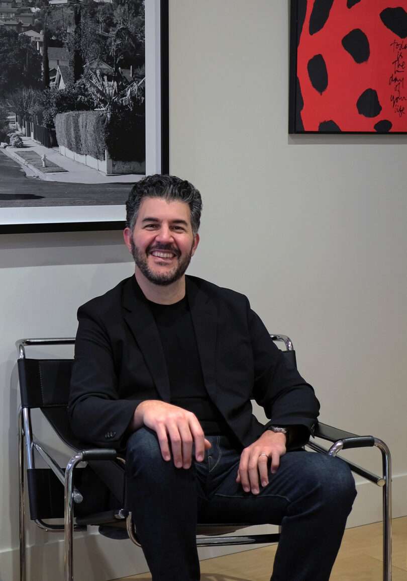 A man with short hair and a beard, wearing a black blazer and jeans, sits smiling in a modern chair in front of two framed artworks on the wall, ready for the Friday 5.