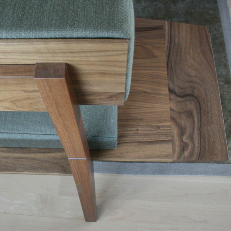 A close-up of a wooden chair leg and armrest, designed by Barry Ginder, upholstered in green fabric, positioned on a wooden surface with visible wood grain.
