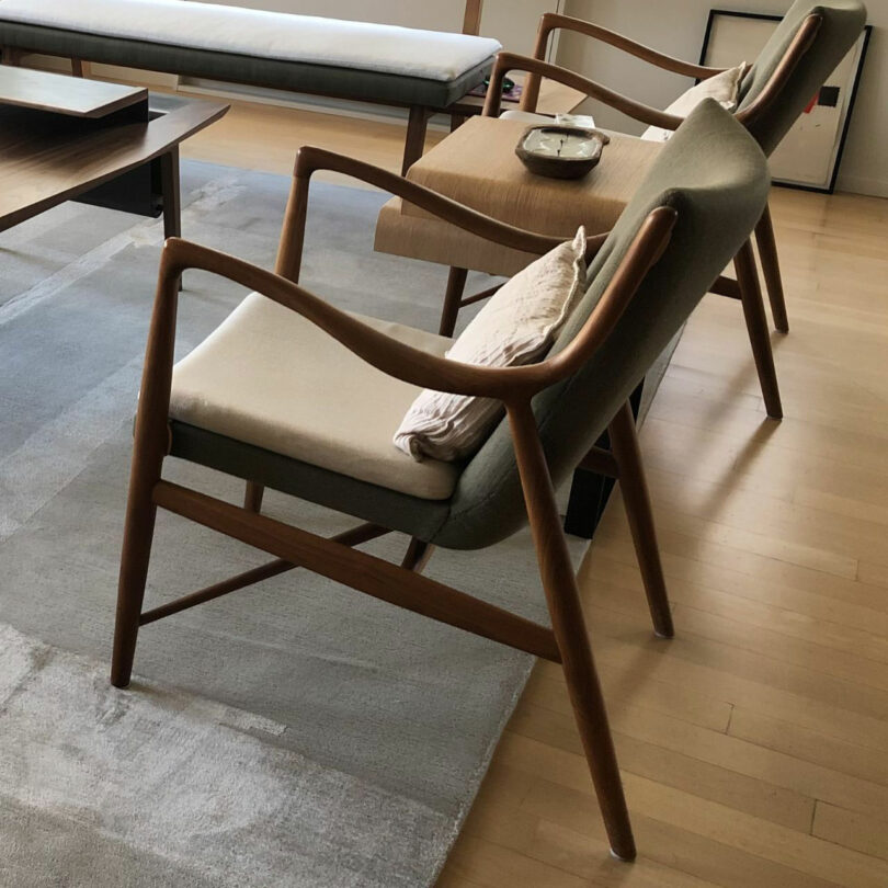 Two modern wooden chairs with gray cushions and beige throw pillows, designed by Barry Ginder, are placed next to a wooden table in a room with light wood flooring and a gray area rug.