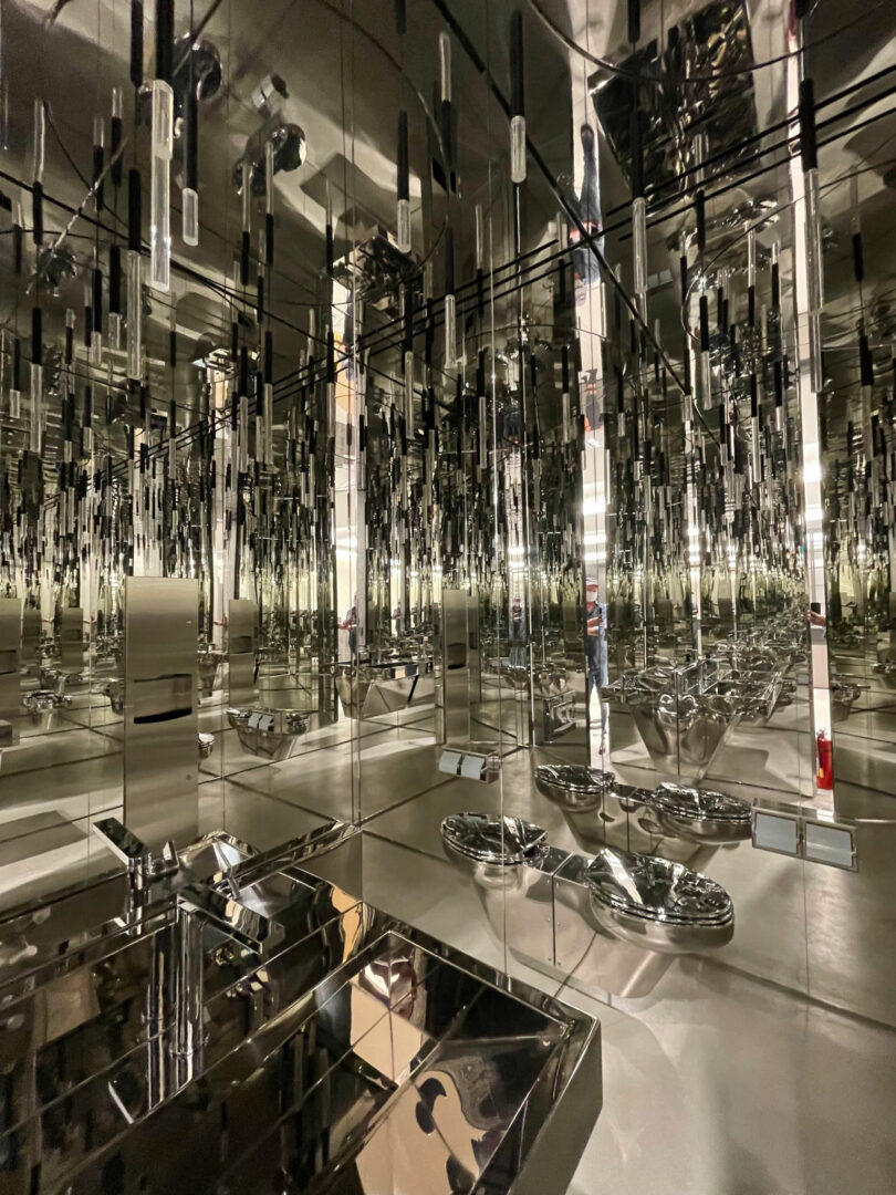 A room filled with mirrors on the walls and ceiling, reflecting multiple sink basins and metallic hanging rods, creates an optical illusion effect reminiscent of a Friday 5 art installation.