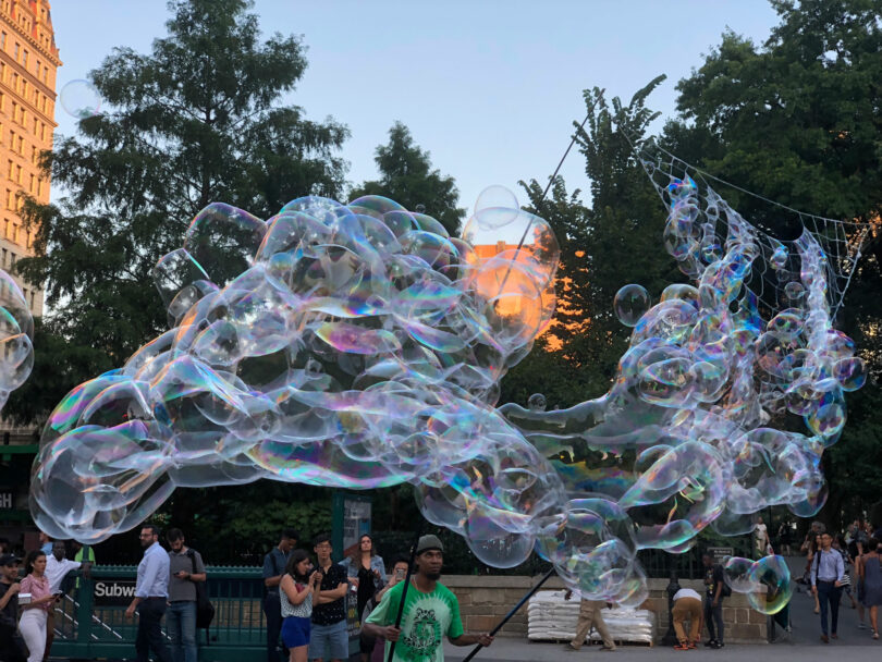 A person creates large soap bubbles in a busy urban park on a Friday 5 evening while people watch and walk by; trees and buildings are visible in the background.