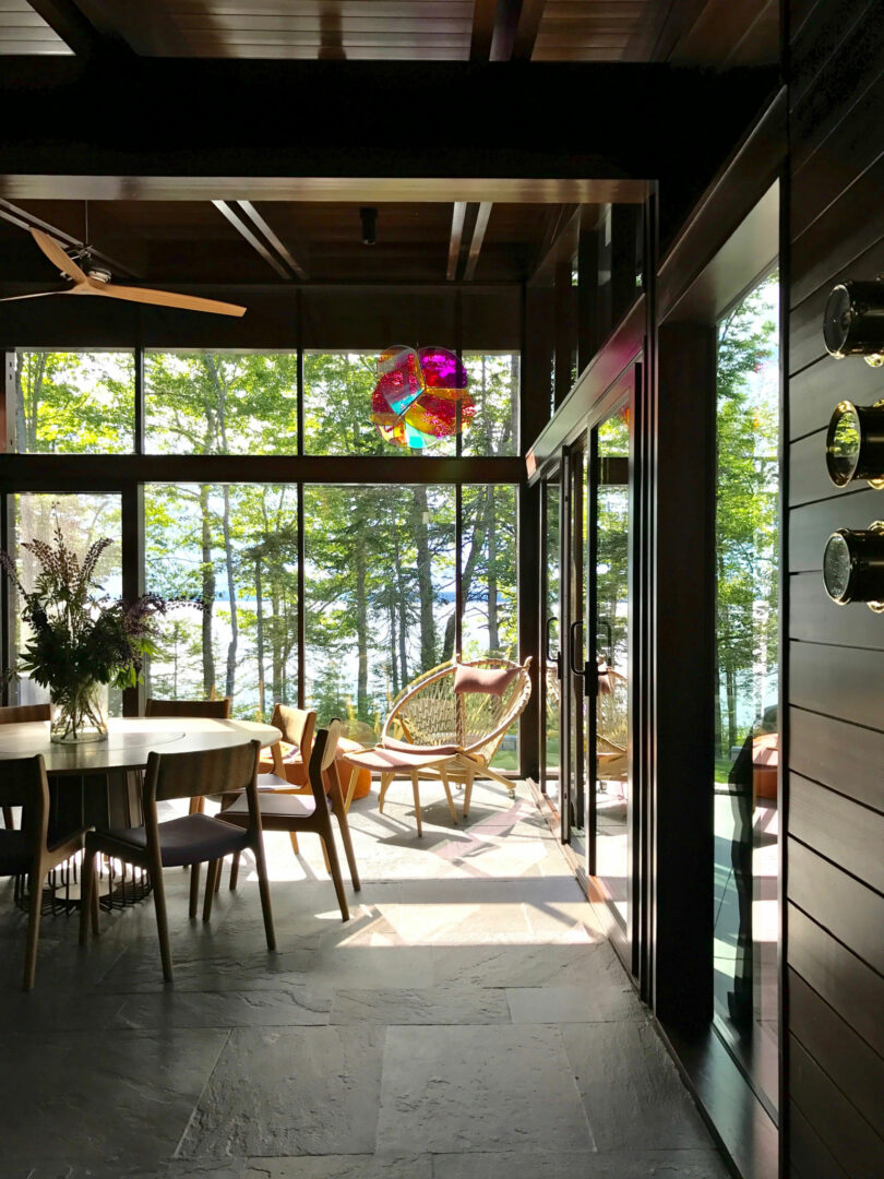 Modern sunlit room with large windows, wooden furniture, and a hanging chair; perfect for relaxing after a Friday 5 run—surrounded by trees with a peaceful lake view in the background.