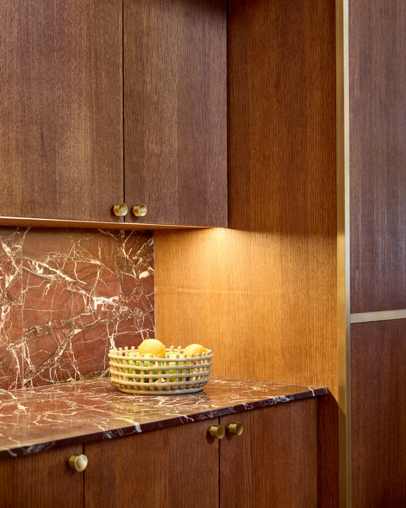A woven basket of lemons and limes from LEGEARD STUDIO sits on a red marble countertop beneath wooden cabinets in a modern kitchen.