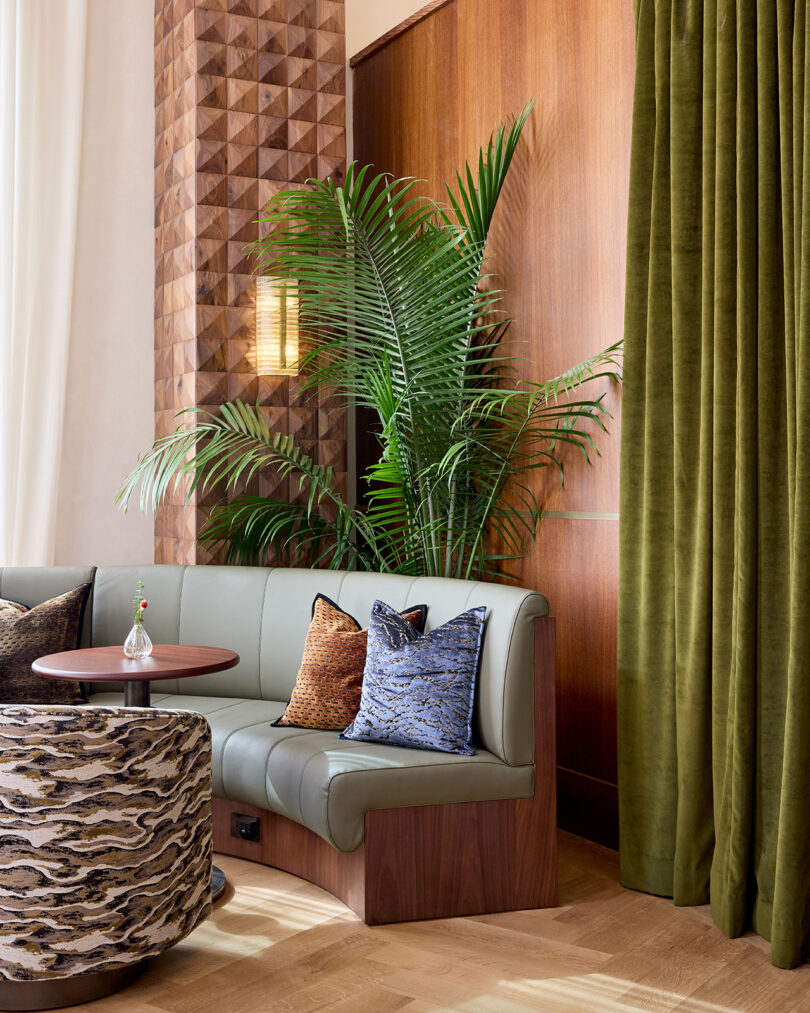 A modern lounge area by LEGEARD STUDIO features a curved green sofa, patterned pillows, a round table, a patterned ottoman, a tall leafy plant, wood paneled walls, and green velvet curtains.