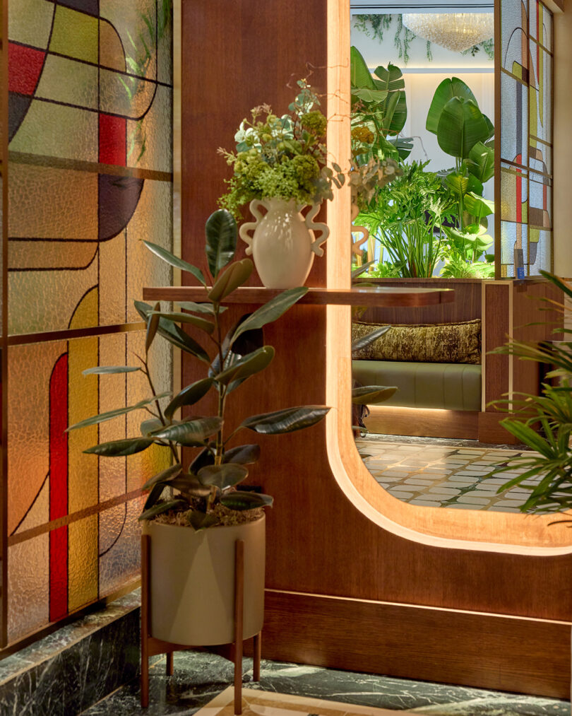 A modern interior by LEGEARD STUDIO features stained glass panels, a potted plant on the floor, a vase with flowers on a shelf, and abundant greenery visible through a window-like opening.
