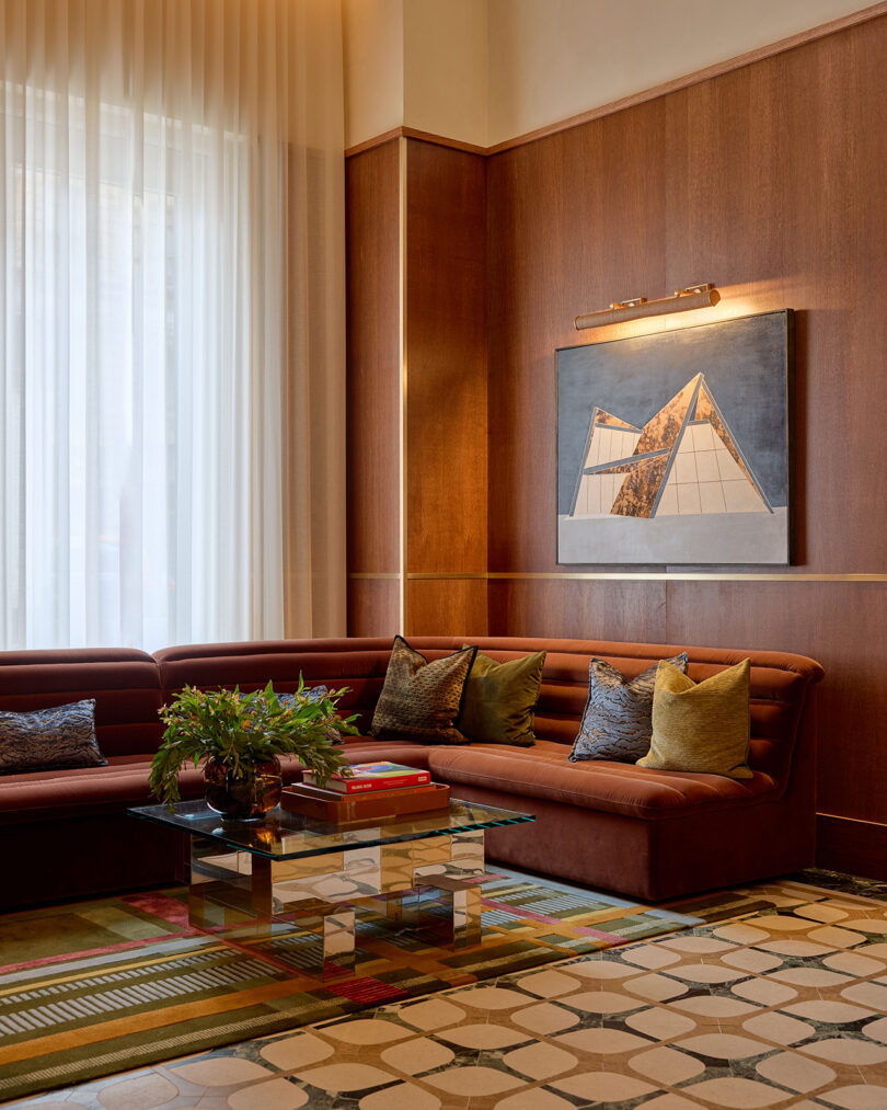 A modern lounge by LEGEARD STUDIO featuring a brown sectional sofa, patterned pillows, a glass coffee table with books, a potted plant, geometric art on a wood-paneled wall, and large sheer curtains.