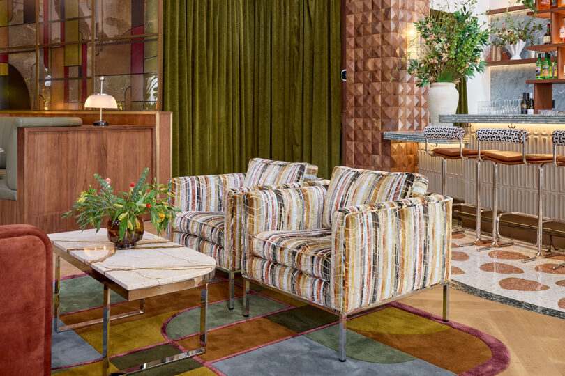 Two patterned armchairs sit by a marble coffee table with a vase of flowers in a modern lounge area featuring green curtains and geometric wall panels, showcasing the sophisticated design style of LEGEARD STUDIO.