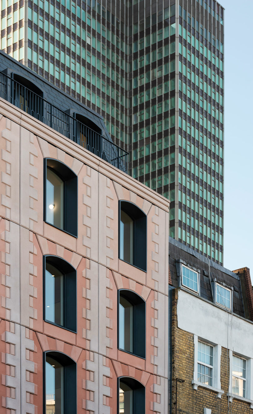 A modern building with pink geometric facade stands in front of a tall glass skyscraper and next to an older brick building with white windows.