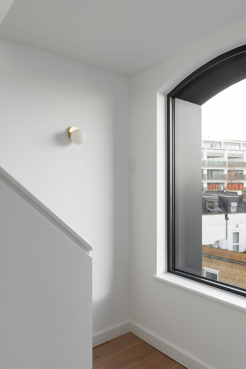 A minimal stairwell with white walls, a modern round wall light, wood flooring, and a large arched window overlooking city buildings.