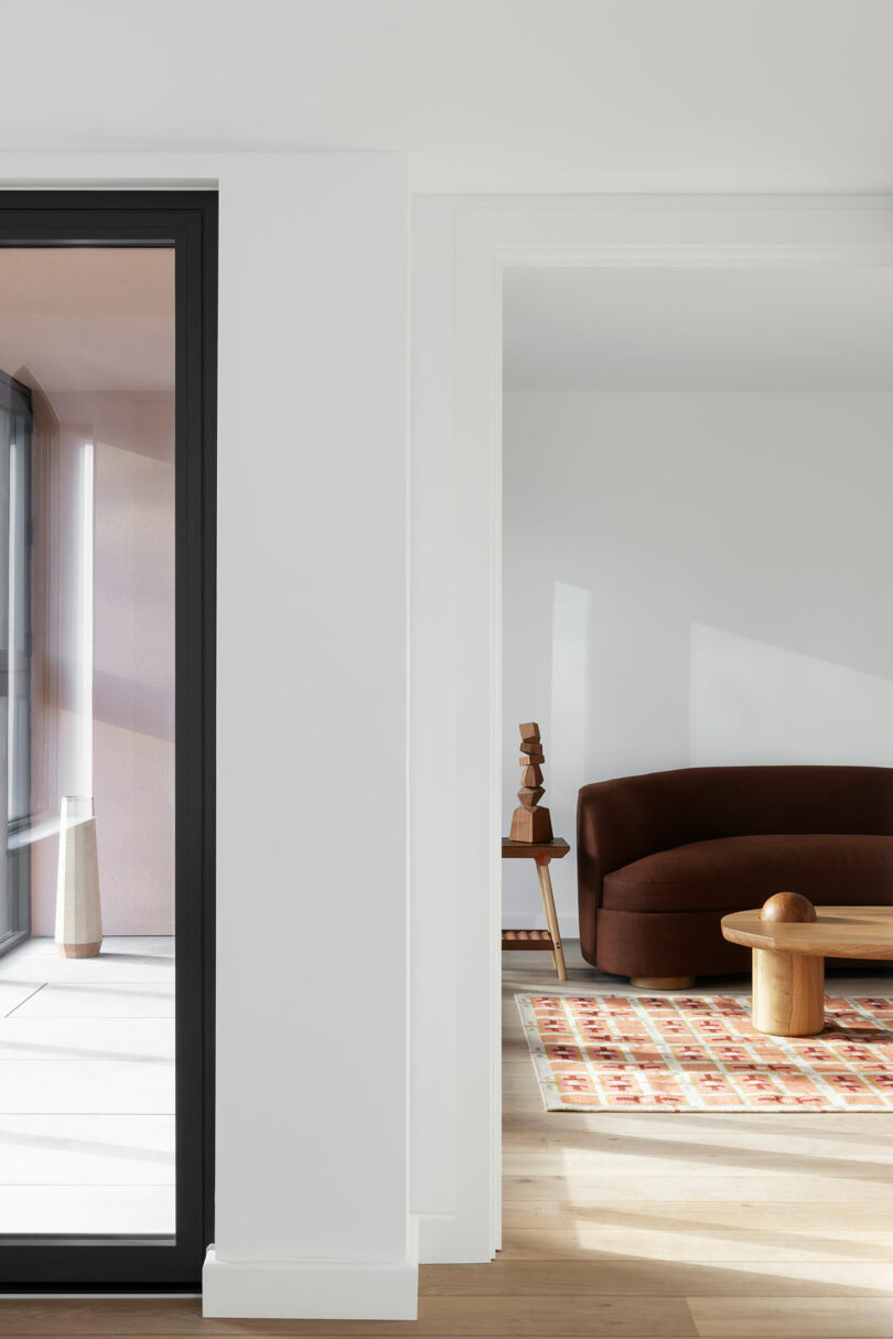 A modern living space with a brown curved sofa, abstract wooden sculptures, a patterned rug, and large windows letting in natural light.