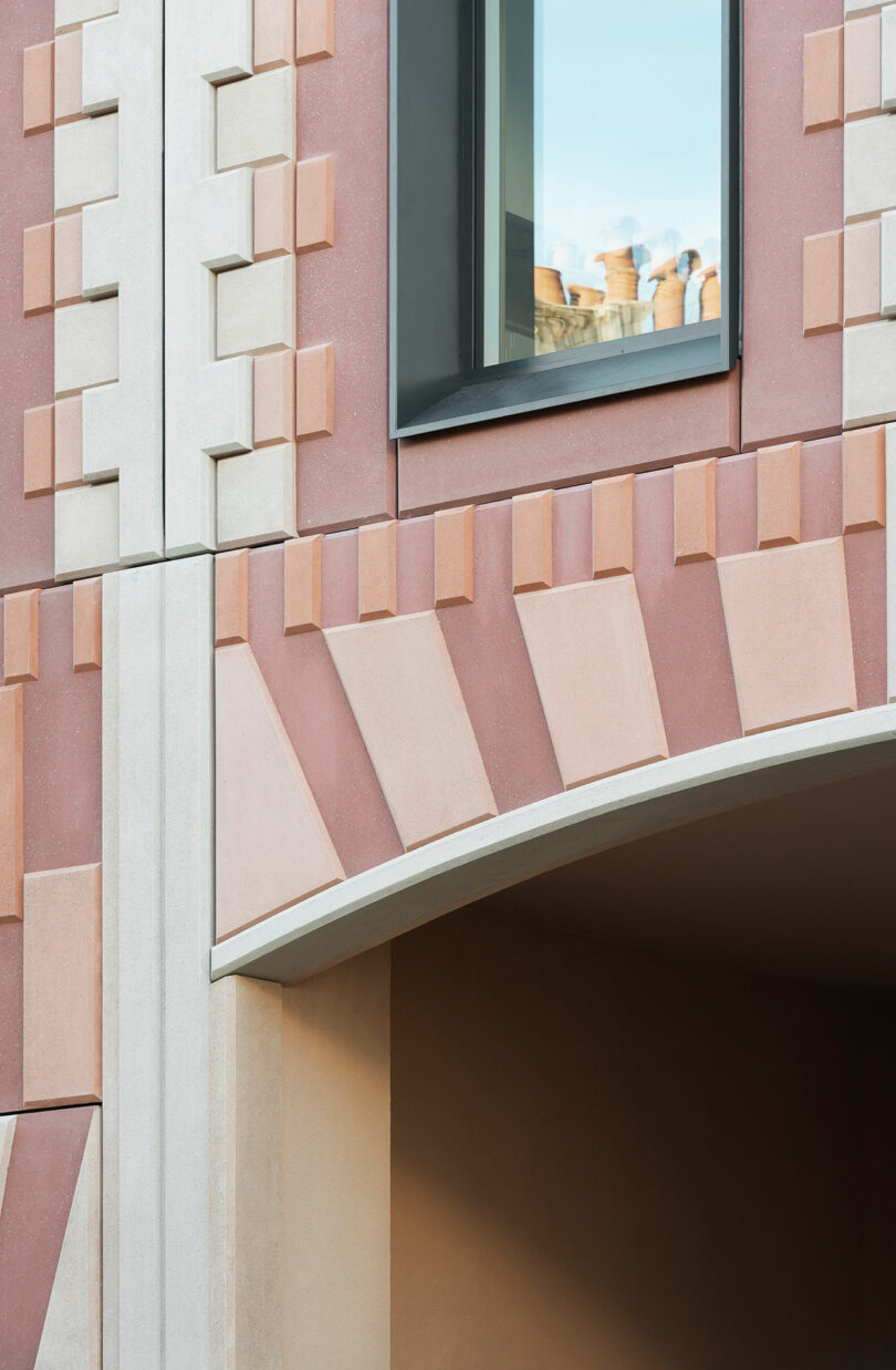 A close-up view of a building facade featuring geometric patterns in pink and cream colors, with a window and an arched passage below.