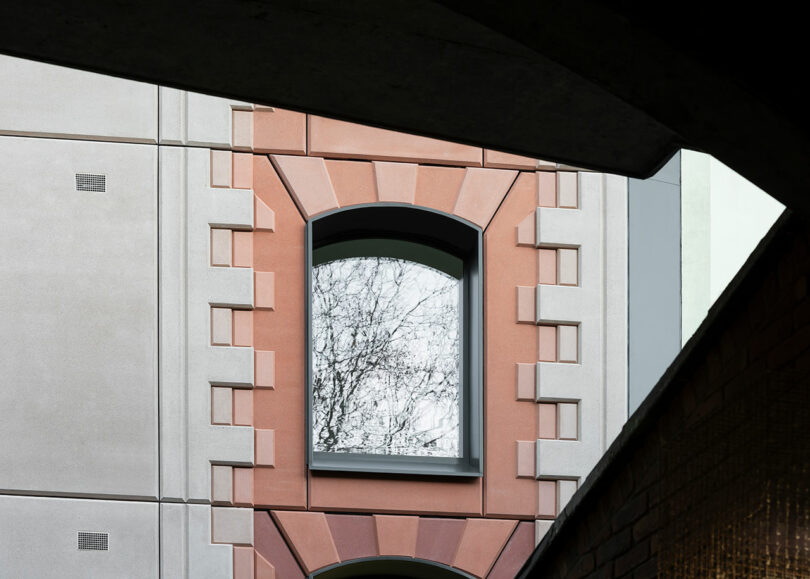 A single arched window with a brick and concrete frame reflects tree branches, viewed through geometric architectural structures.