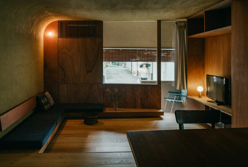 A minimalist living room with wood paneling, a built-in bench, a TV on a shelf, a window with bamboo blinds, and a chair near a small table with flowers.