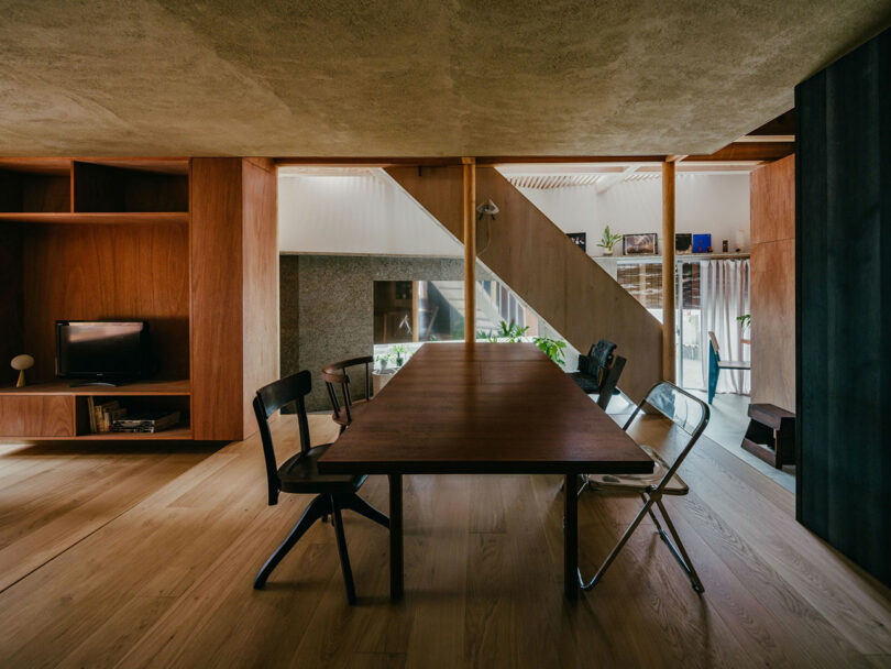 Modern dining area with a large wooden table, mixed chairs, wooden flooring, and open shelving, connected to a living space with natural light and contemporary decor.