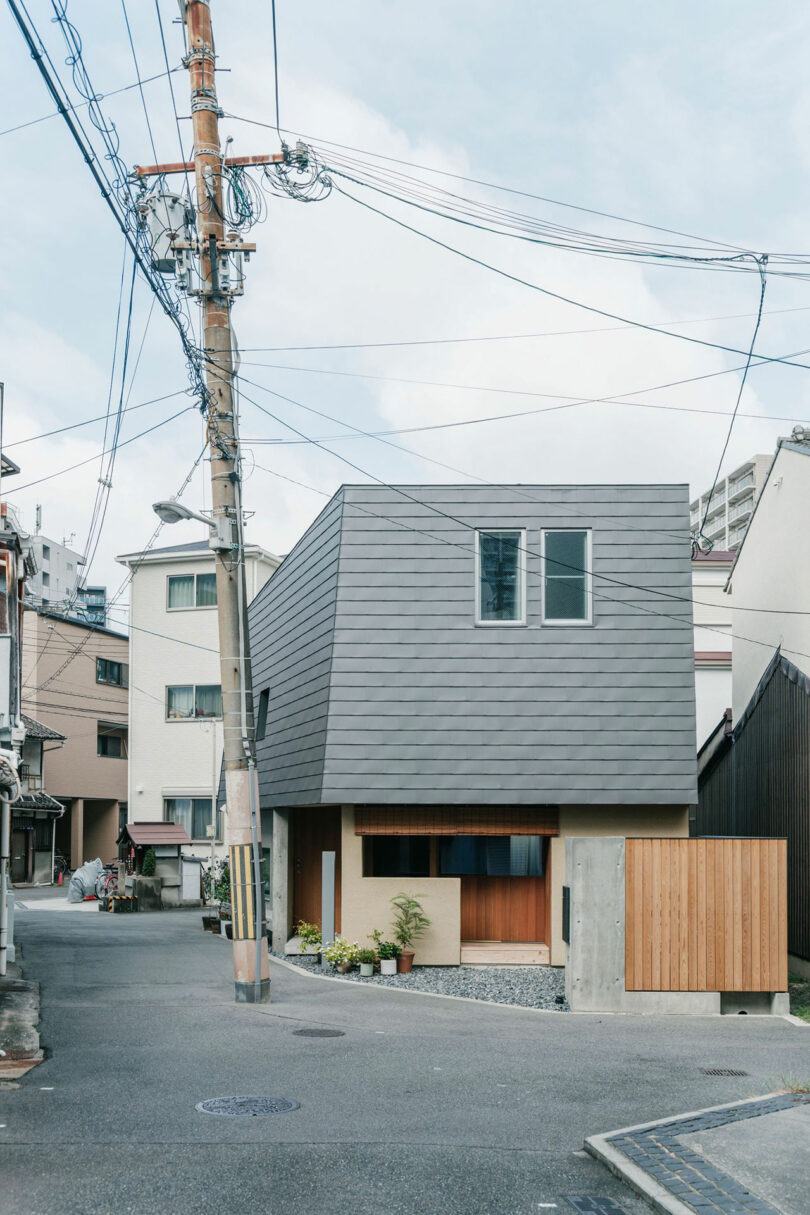 A modern, angular house with a gray sloped roof sits at a street corner beside a wooden utility pole with overhead wires in an urban neighborhood.