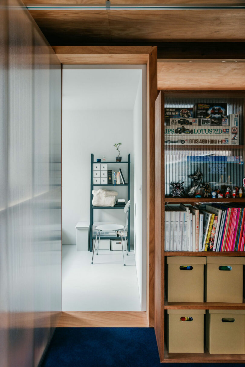 A view through a wooden-framed doorway into a minimalist white room with a desk, chair, shelving unit, and organized storage boxes on the right.