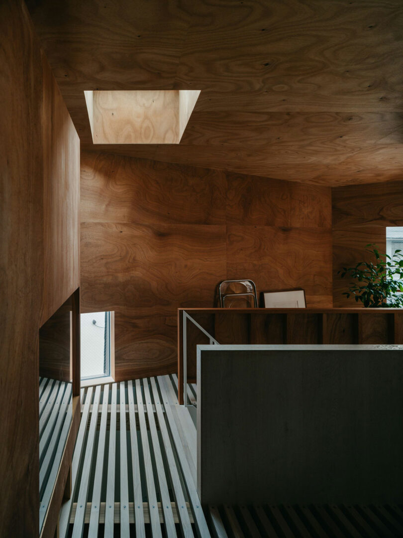 Interior space with wooden walls and ceiling, a skylight, a metal slatted floor, a white railing, and a window letting in natural light. A plant and a folded ladder are visible in the corner.