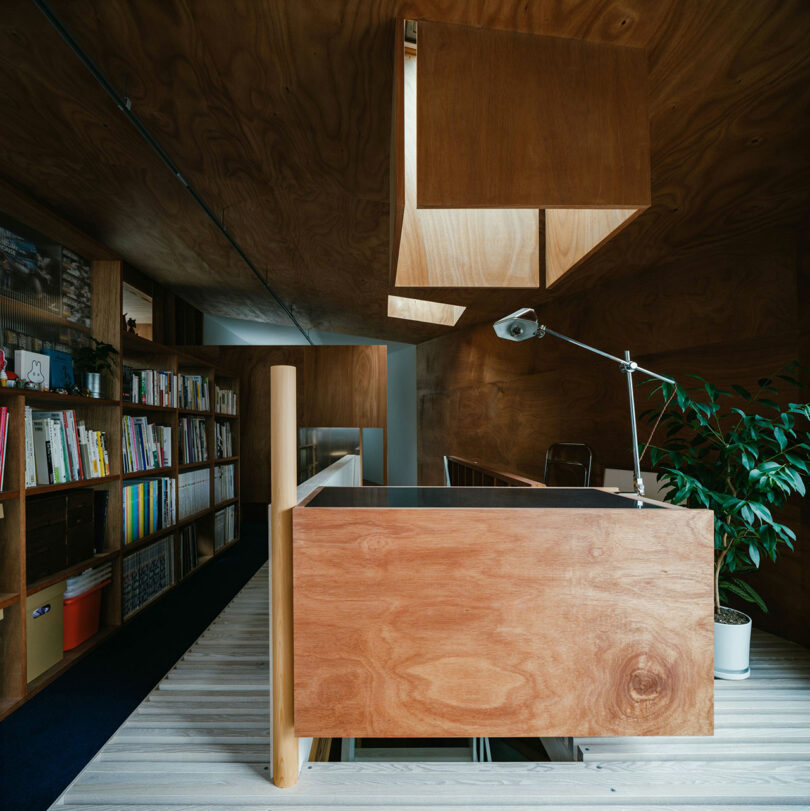 Modern office interior with wood paneling, a built-in desk, a desk lamp, bookshelves, and a potted plant. Natural light comes through an opening in the ceiling.