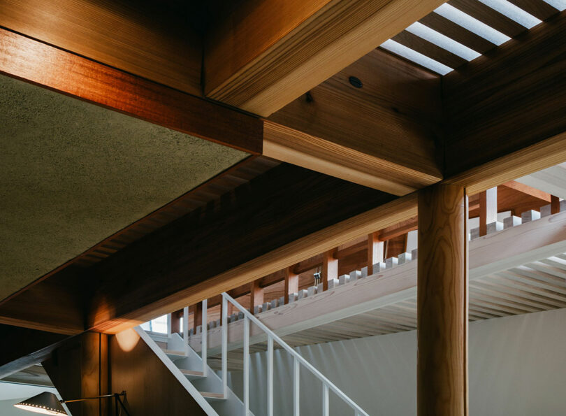 Interior view of a modern building featuring exposed wooden beams, a partial staircase with a white railing, and natural light filtering through slatted ceiling panels.