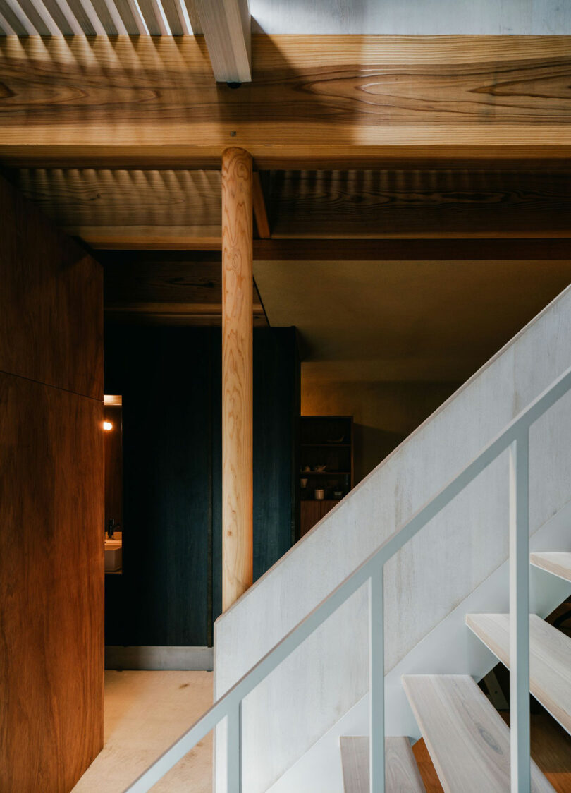 Modern interior with light wood beams, a central wood column, white staircase, and a dark wall with a small lit room visible in the background.