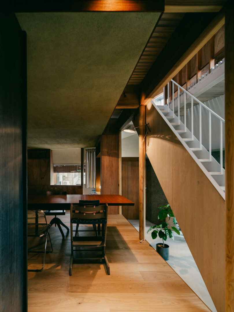 Modern interior with wooden floors and walls, a dining table with chairs, a staircase with white railing, and a potted plant beneath the stairs. Natural light enters through windows.