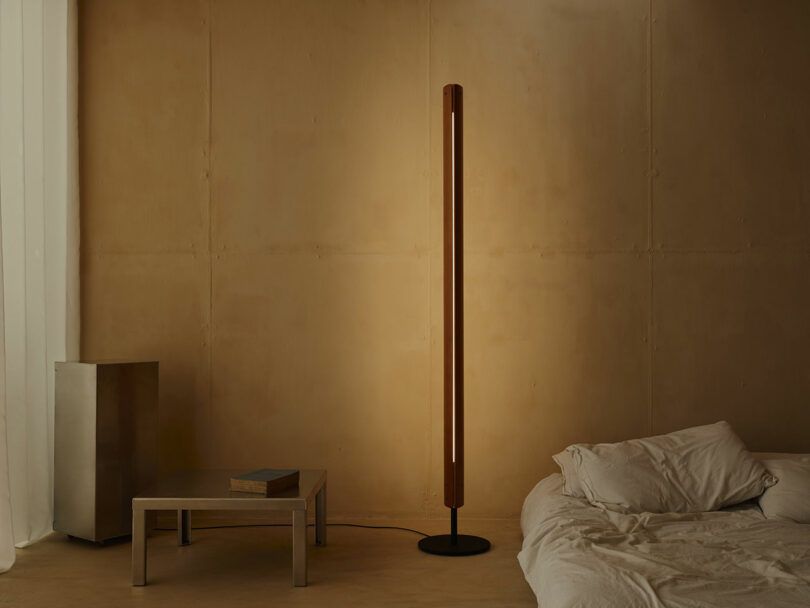 A tall, slim floor lamp emits warm light beside an unmade bed, metal side table, and square metallic cabinet in a minimal beige room with sheer curtains.