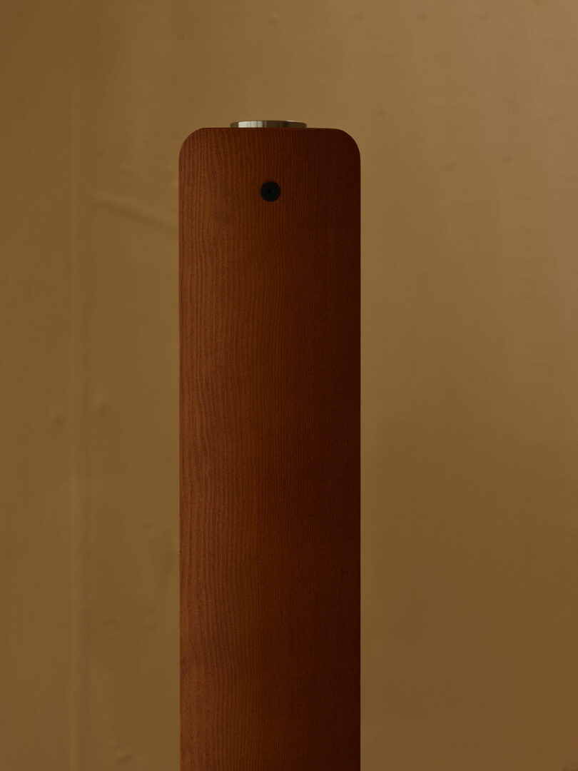 A tall, rectangular wooden post with rounded edges, featuring a small black circular detail and a metallic button on top, set against a beige background.