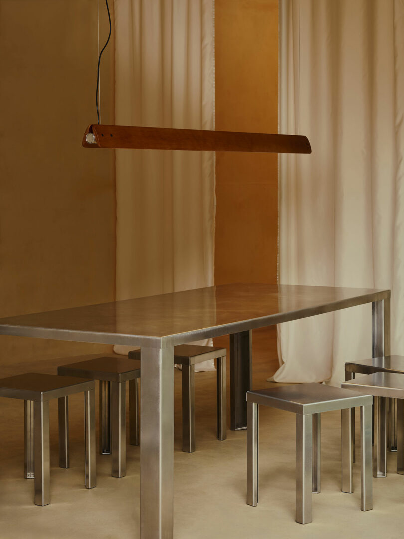 A minimalist dining area with a rectangular metal table, four matching metal stools, a wooden pendant light, and beige curtains in the background.