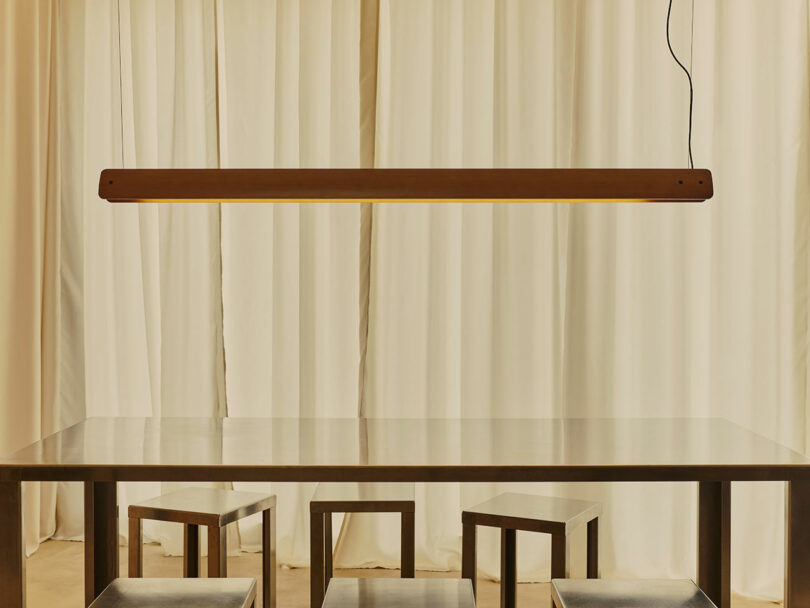A rectangular pendant light hangs above a wooden table with four matching stools, set against cream-colored curtains.