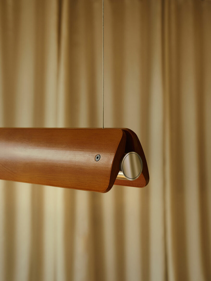 A wooden pendant light fixture with a cylindrical metal accent hangs from a thin wire in front of beige curtains.