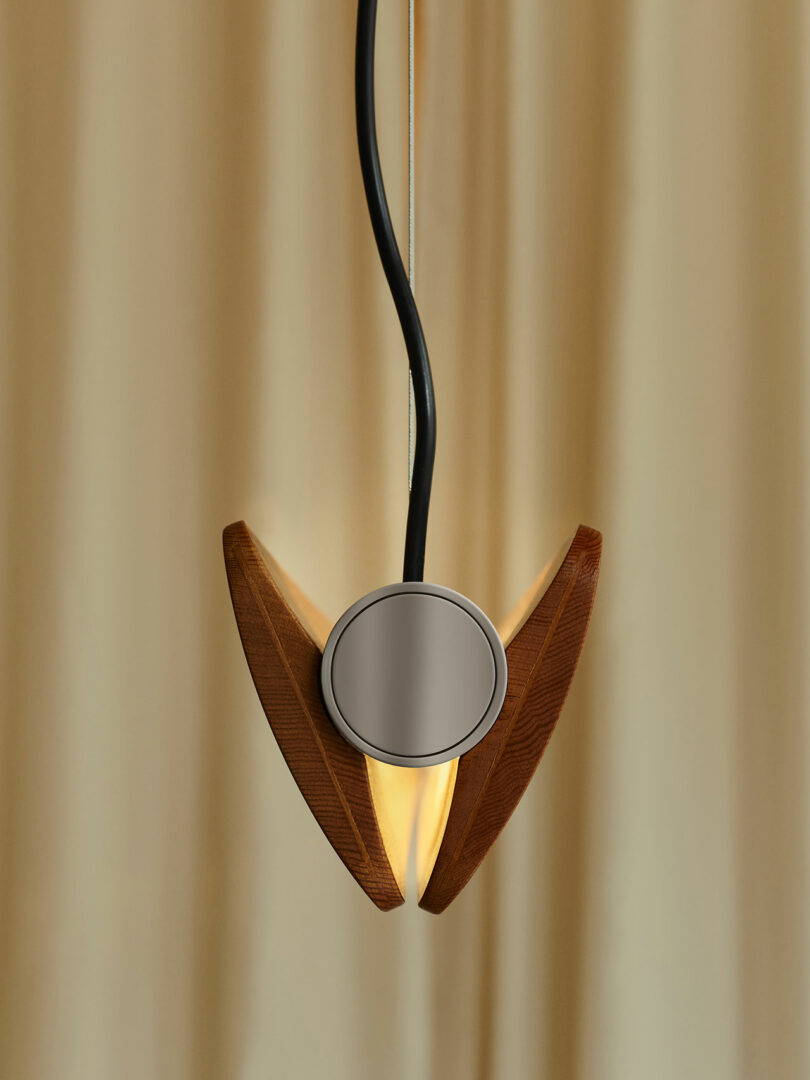 Pendant light with a wooden fixture and a round central metallic element, suspended by a black cord against a beige curtain background.