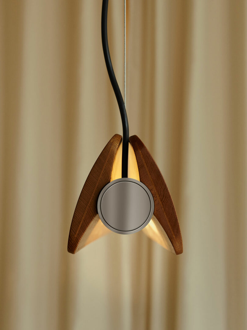 A modern pendant light with a round metal center and three wooden panels, suspended from a black cord against a beige curtain background.