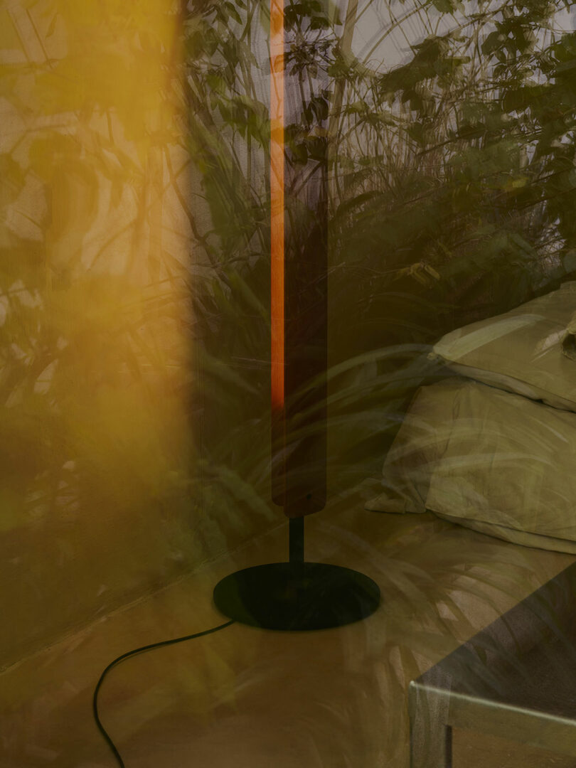 A floor lamp with a black base stands next to a bed and a side table, with leafy plant shadows cast on the wall and bedding.