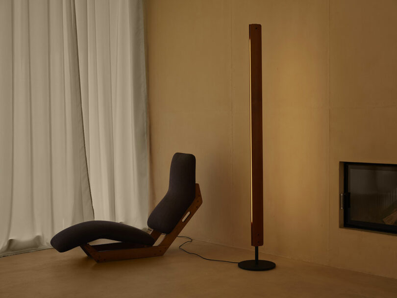 A modern lounge chair with a wooden frame and dark cushion sits beside a tall, vertical floor lamp in a minimalist room with beige walls and a white curtain.
