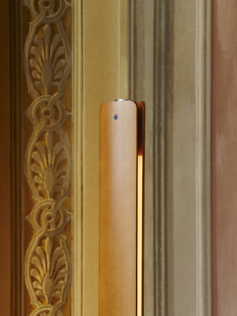 A vertical wooden or metal light fixture stands in front of an ornate, decorative wall with intricate patterns and muted colors.