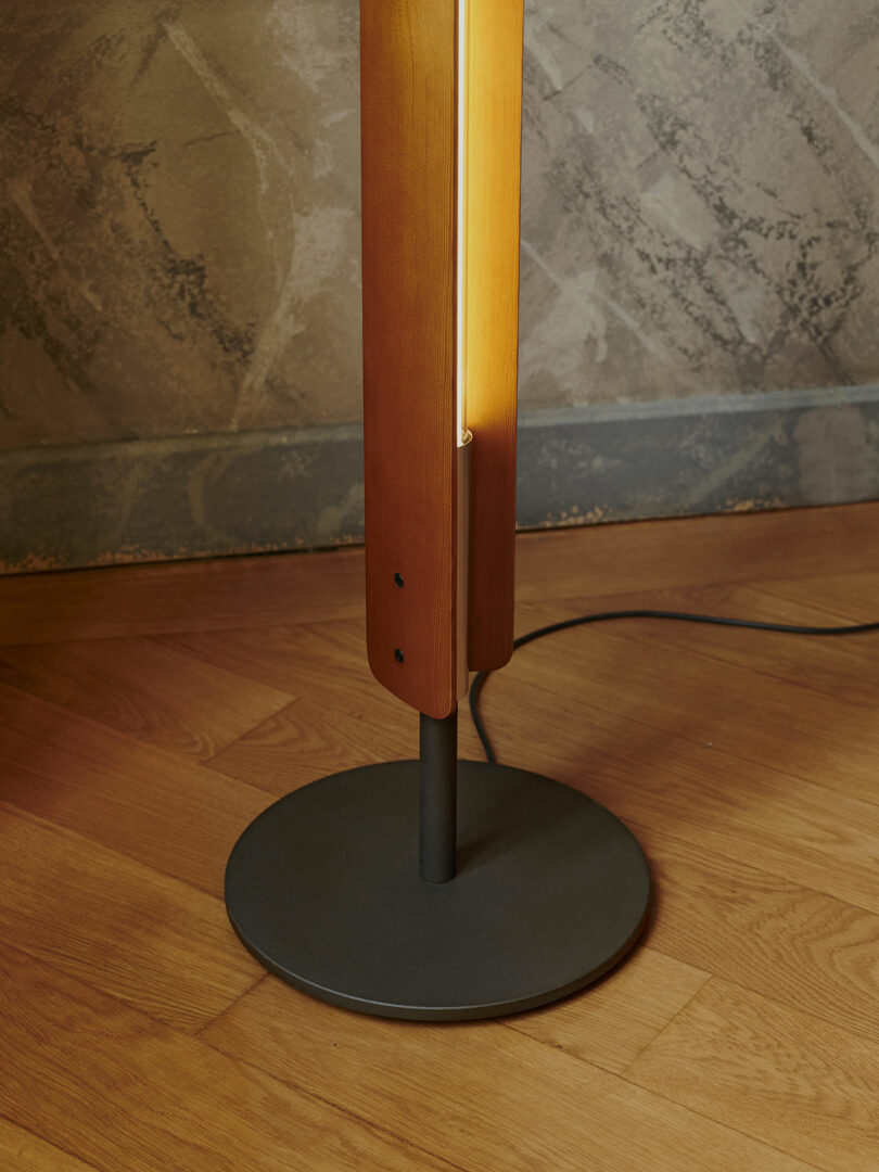 A close-up of a floor lamp with a wooden vertical body, metal base, and cord on a wooden floor against a textured wall.