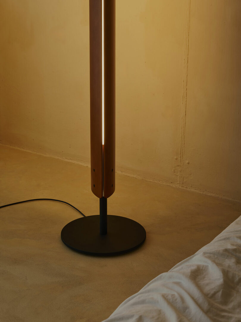 A wooden floor lamp with a vertical light strip stands on a round black base next to a bed with a white blanket in a beige room.