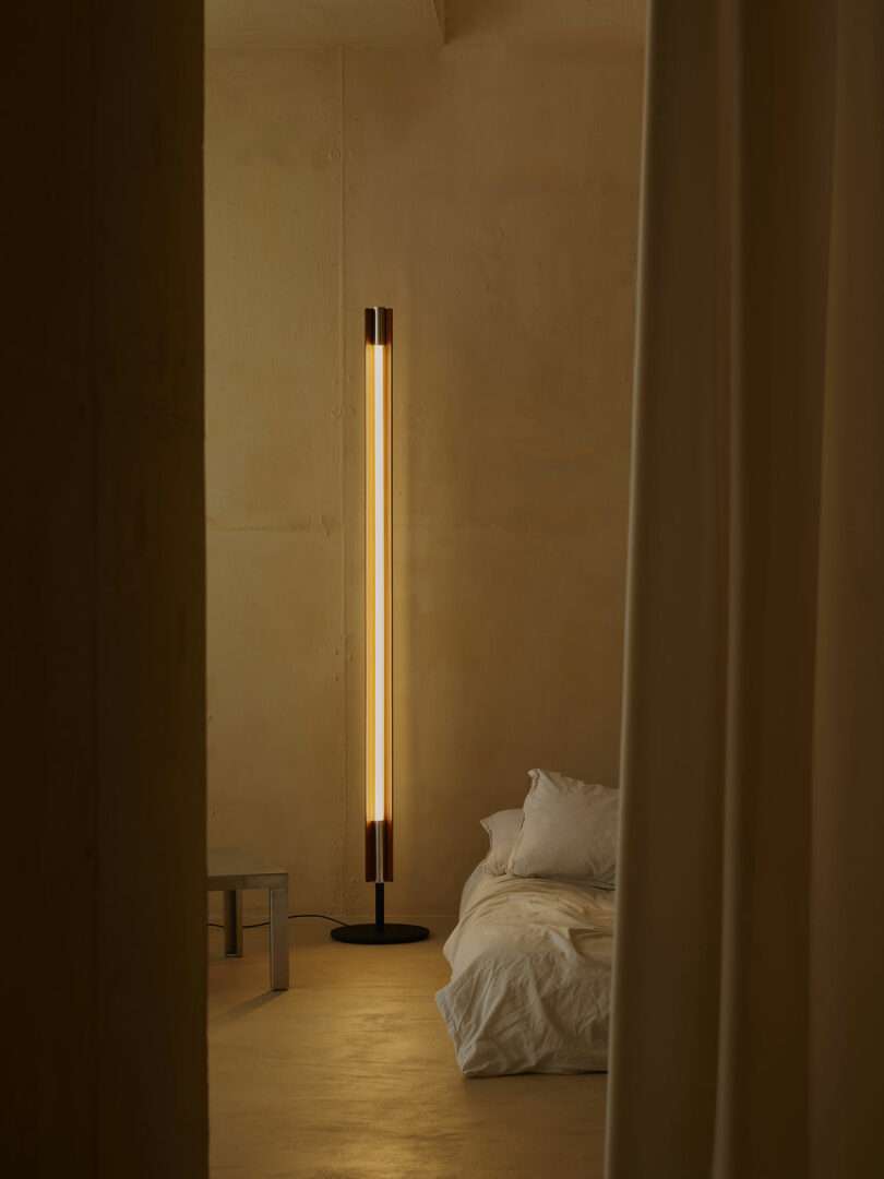 A minimalist bedroom with beige walls, a low bed with white bedding, a vertical floor lamp emitting warm light, a metal side table, and cream-colored curtains.