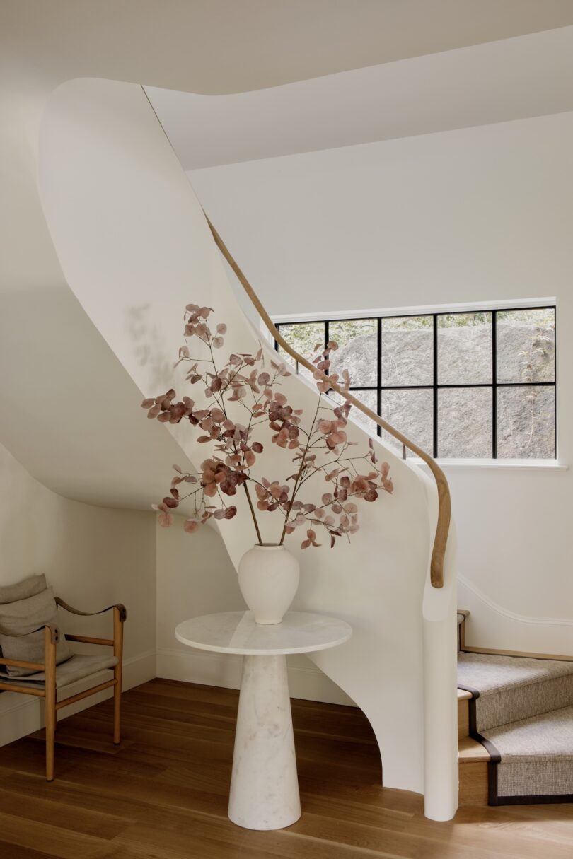 A marble table with a white vase of pinkish branches stands in front of a curved white staircase and a window with black frames in a modern interior.