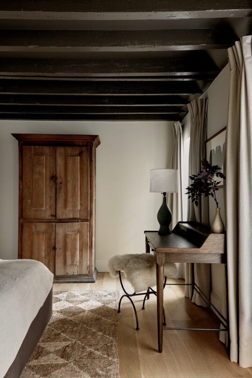 A cozy bedroom with a wooden armoire, a writing desk with a lamp and vase, a fur-covered stool, and beige curtains. Exposed dark ceiling beams and natural light are present.