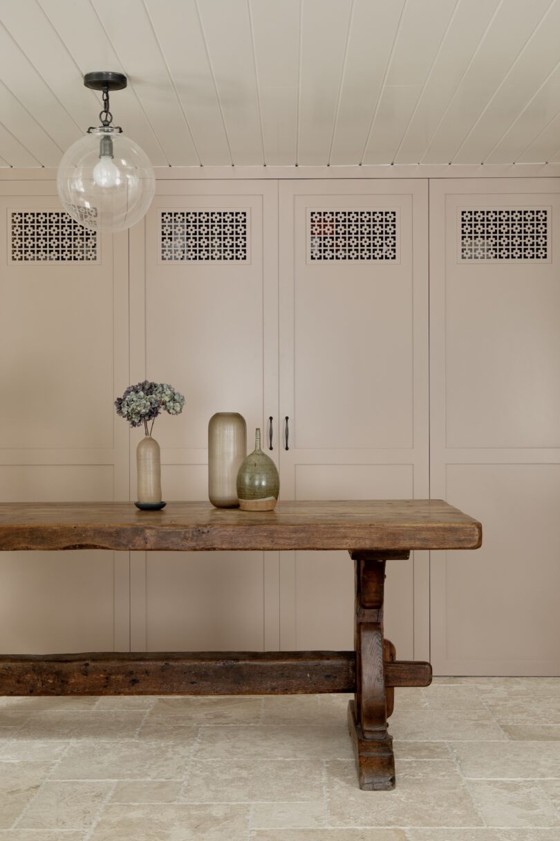 A rustic wooden table with three ceramic vases and dried flowers stands in front of beige cabinets with decorative vent panels; a glass pendant light hangs above.