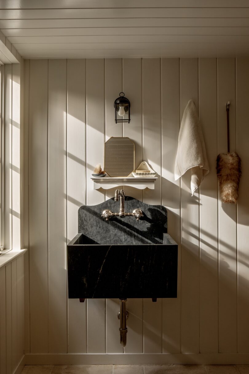 A small bathroom with a black marble sink, towel hanging on a hook, wall-mounted shelf with toiletries, and natural light streaming through a window.