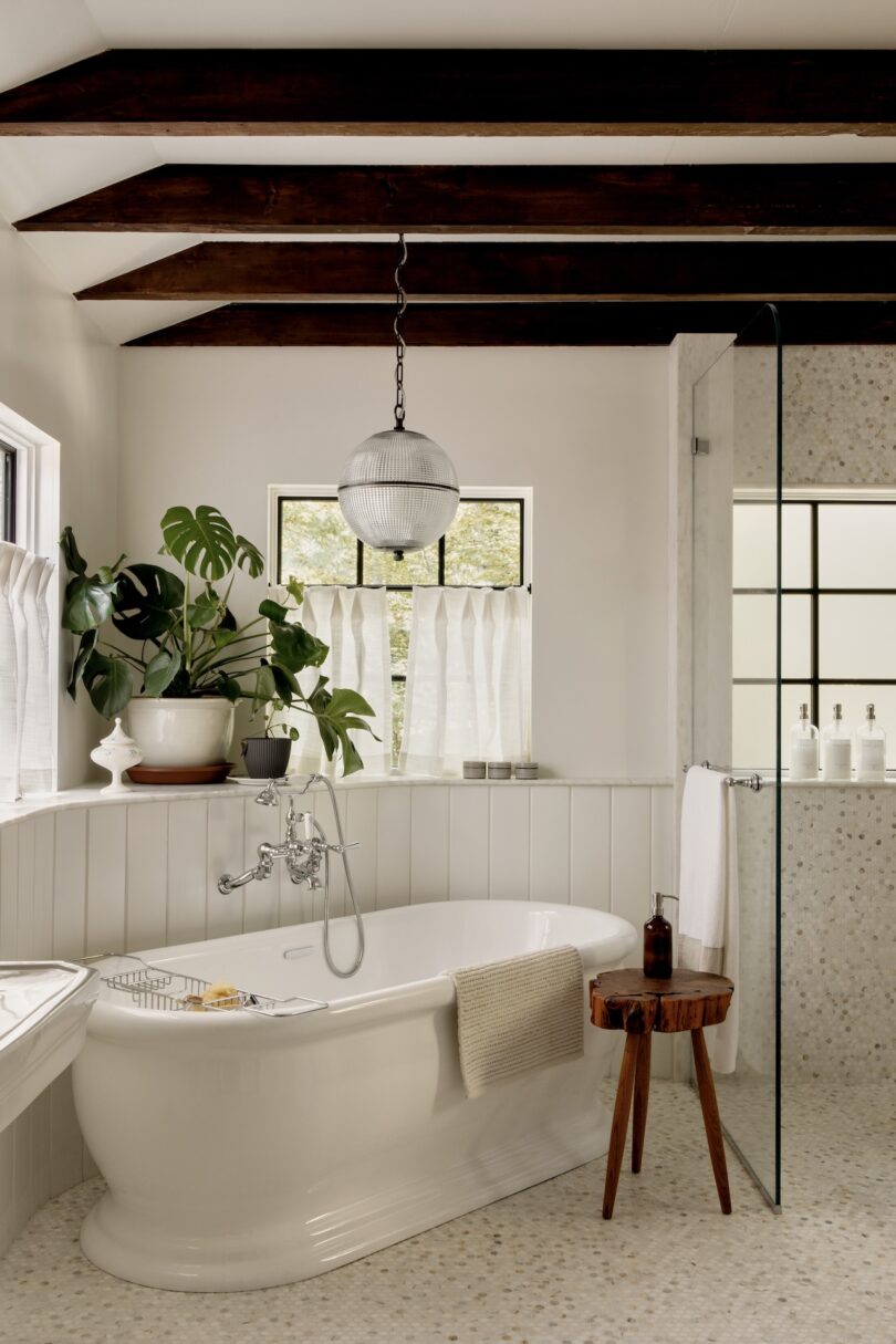 A white bathroom features a freestanding tub, a wooden stool, potted plants, exposed dark beams, and a glass shower enclosure with natural light from windows.