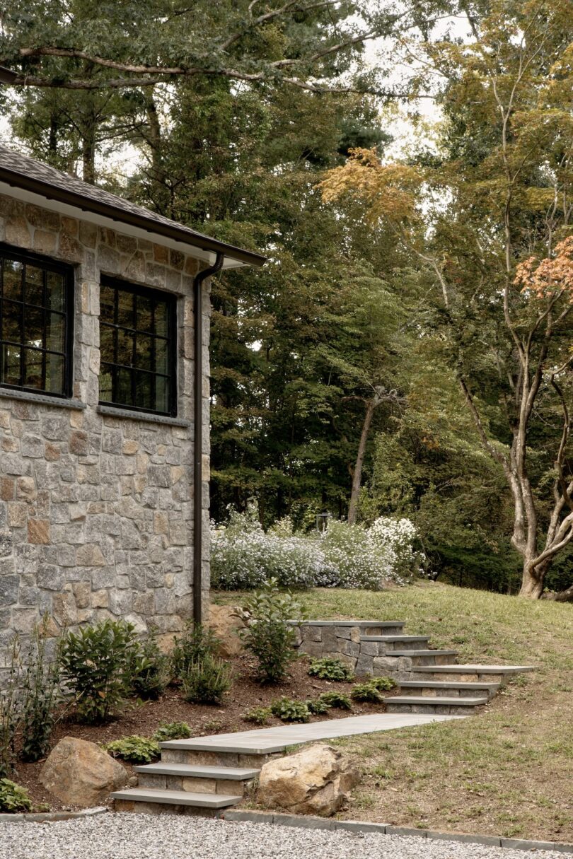 Stone steps lead up a landscaped slope beside a stone building, with shrubs, flowering plants, and tall trees in the background.